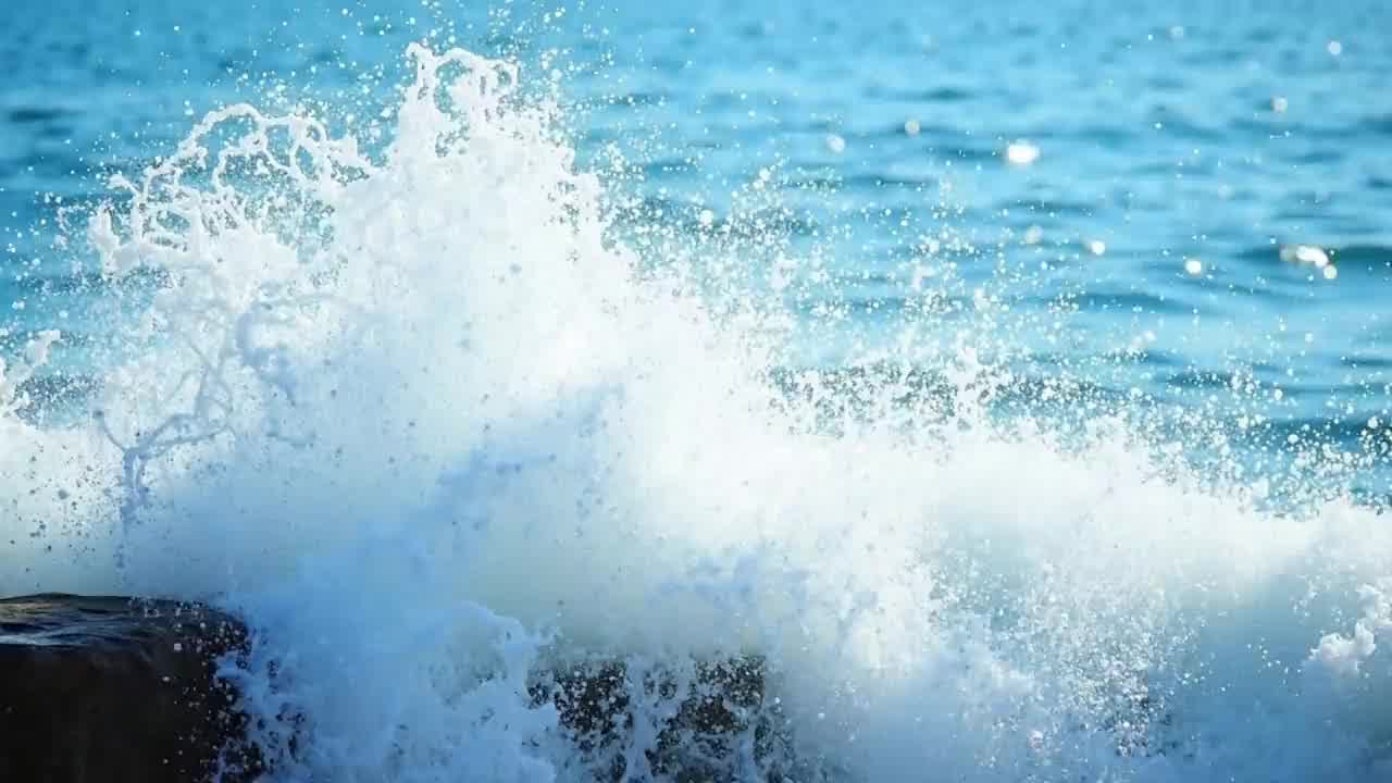 A wave bursts into bright white spray over a stone breakwater in strong sunshine