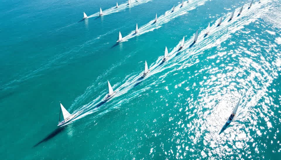 Aerial photograph of a sailboat fleet crossing a sunlit turquoise bay, each boat trailing a thin wake